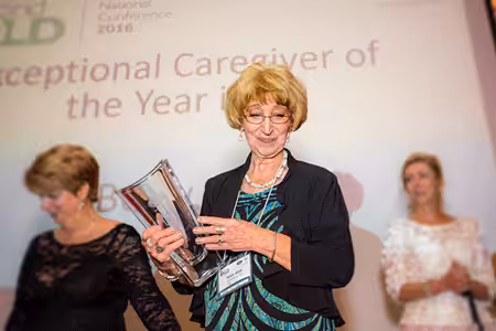 A woman holding a glass award stands in front of a screen that reads Exceptional Caregiver of the Year at a conference, with two other women standing nearby in the background.