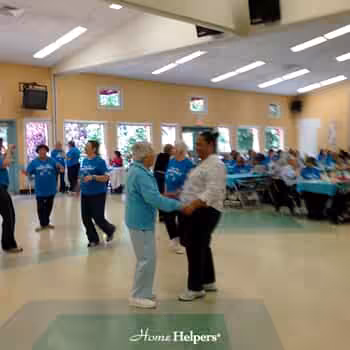 A group of seniors gathers in a large, bright room; some are dancing in pairs while others sit at tables. Most people wear matching blue shirts. The atmosphere appears lively and social. Home Helpers logo is visible.