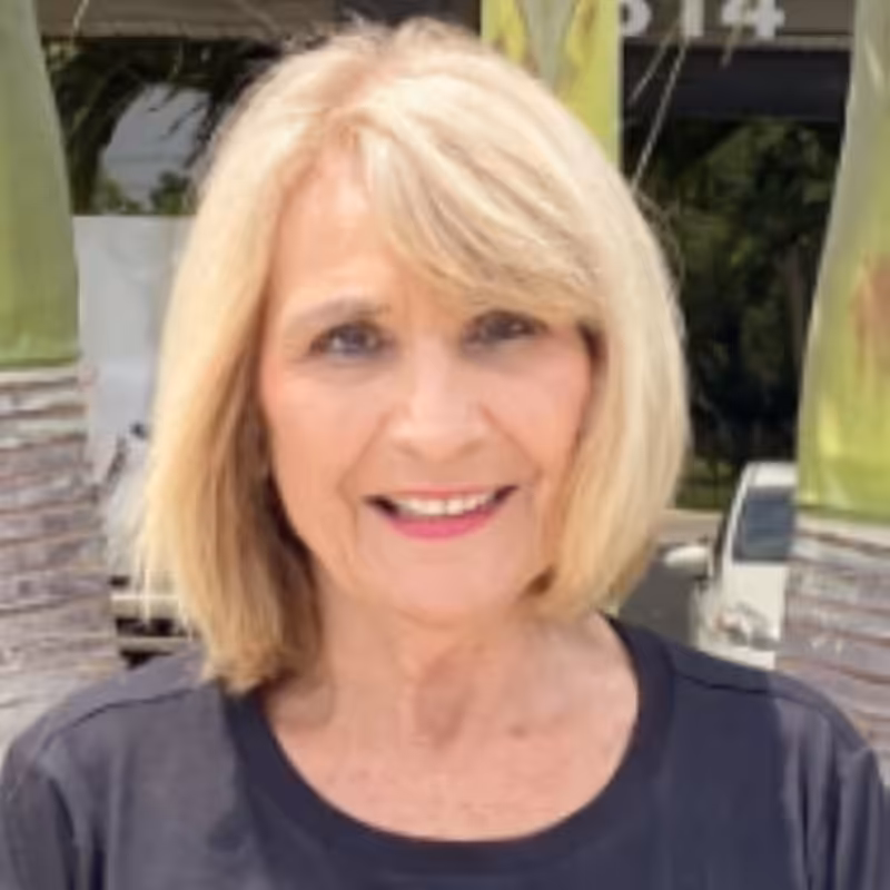 A smiling older woman with blonde hair stands outdoors in front of palm trees and a building, wearing a black shirt.