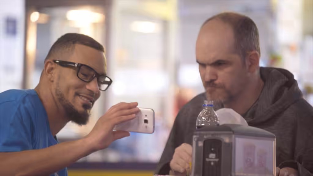 Two men sitting at a table, one smiling and holding up a smartphone to show something to the other, who looks focused and serious. There are food items and a water bottle on the table.