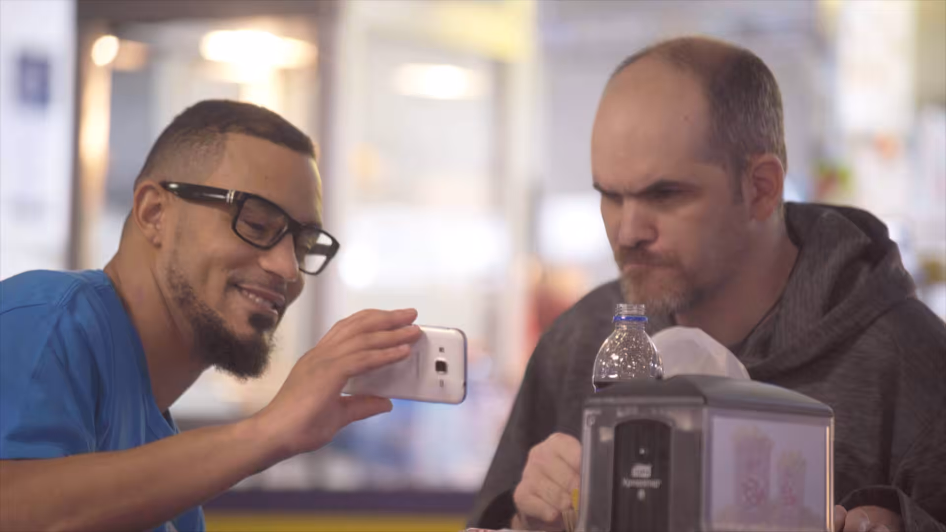 Two men sitting at a table, one smiling and holding up a smartphone to show something to the other, who looks focused and serious. There are food items and a water bottle on the table.
