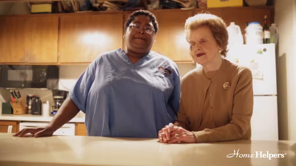 A caregiver in blue scrubs stands beside an elderly woman in a tan sweater, holding her hand at a kitchen counter. The Home Helpers logo appears in the bottom right corner.
