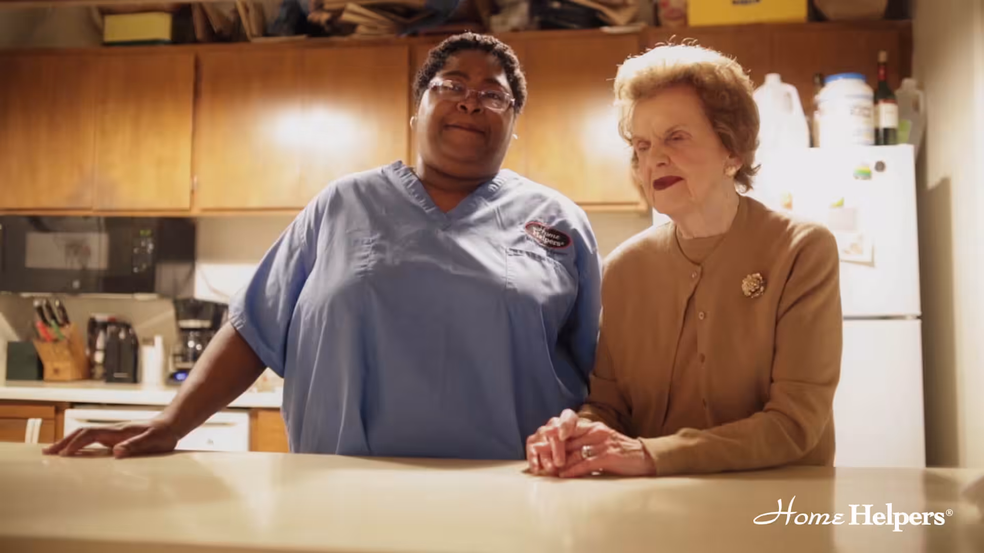 A caregiver in blue scrubs stands beside an elderly woman in a tan sweater, holding her hand at a kitchen counter. The Home Helpers logo appears in the bottom right corner.