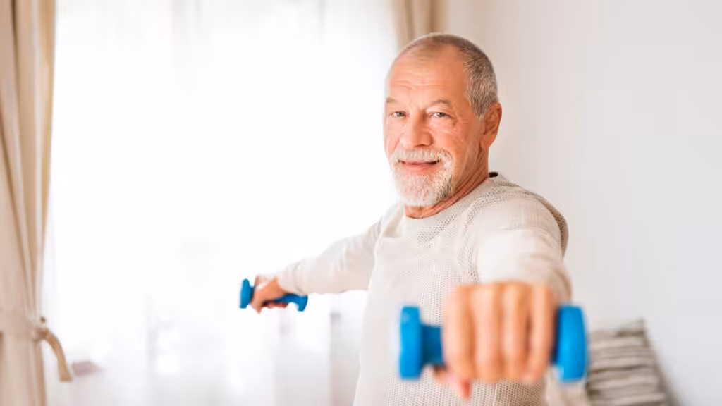 An older man with a gray beard smiles while exercising at home, holding blue dumbbells with arms extended to the sides. Bright window light fills the room in the background.
