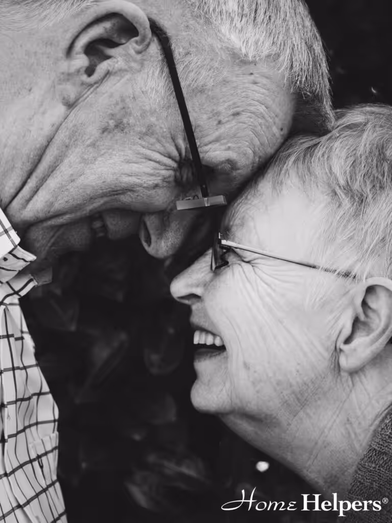 An elderly couple touches foreheads and smiles warmly at each other. Both wear glasses, and the woman is laughing. The image is in black and white with Home Helpers text at the bottom right.
