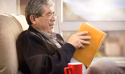An older man with gray hair and glasses sits comfortably in a chair, reading a book with a yellow cover. A red mug rests on the armrest beside him, and sunlight streams through a window in the background.