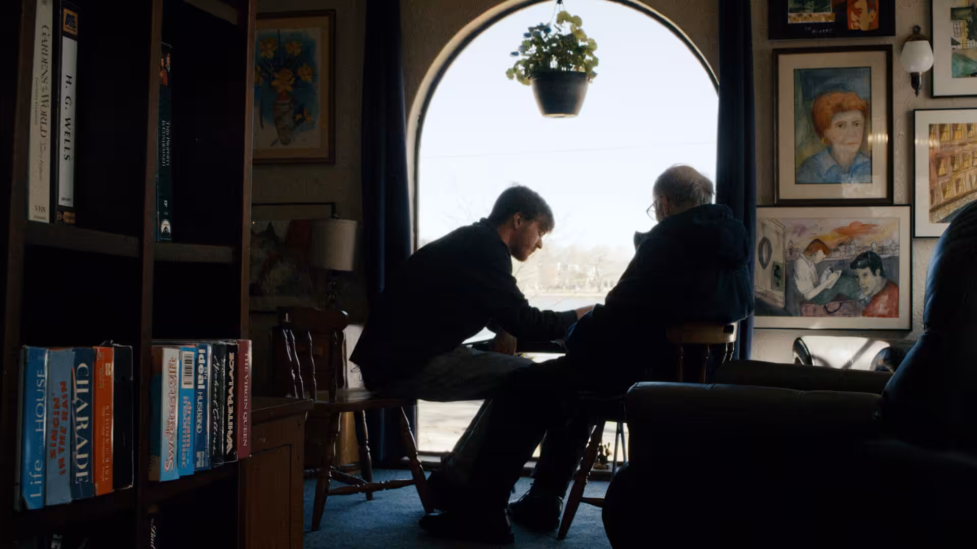 Two people sit facing each other in a cozy, art-filled room with a large arched window. One person leans forward, holding the others hand, while sunlight streams in, illuminating bookshelves and framed portraits on the walls.
