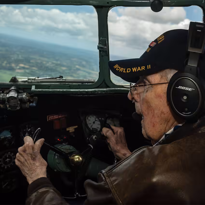 An elderly man wearing a headset and a World War II cap pilots an aircraft, holding the controls and looking out the cockpit window at the landscape below.