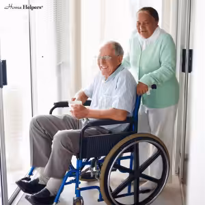 An elderly woman stands behind and smiles at an elderly man in a wheelchair, who is also smiling. They are indoors near large windows, and the man holds a white mug. The logo Home Helpers is in the top left corner.