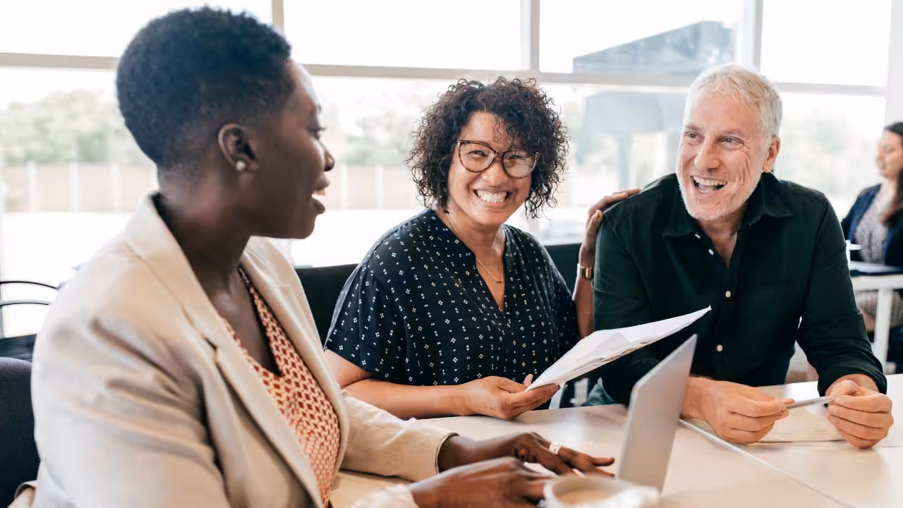 Three adults sit at a table in a bright office, smiling and talking. One woman holds papers, another woman speaks, and a man is holding a document. A laptop and papers are on the table.