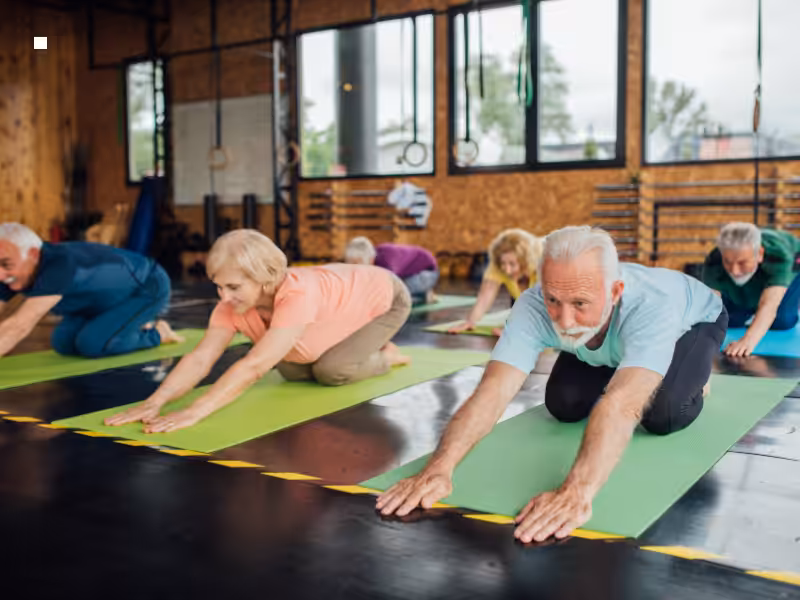 A group of older adults practice yoga on mats in a gym, stretching forward in a childs pose position. The room has large windows and exercise equipment in the background.