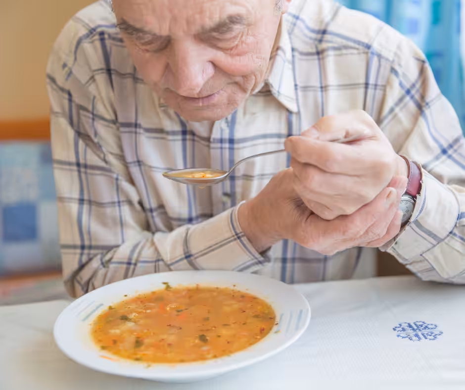 An elderly man wearing a plaid shirt sits at a table, holding a spoonful of soup above a white bowl filled with chunky vegetable soup. He appears to be looking closely at the spoon.