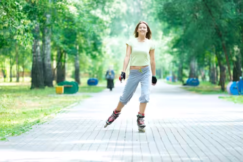 A young woman in casual clothes rollerblades on a paved path in a green, tree-filled park on a sunny day, smiling with arms relaxed by her sides.