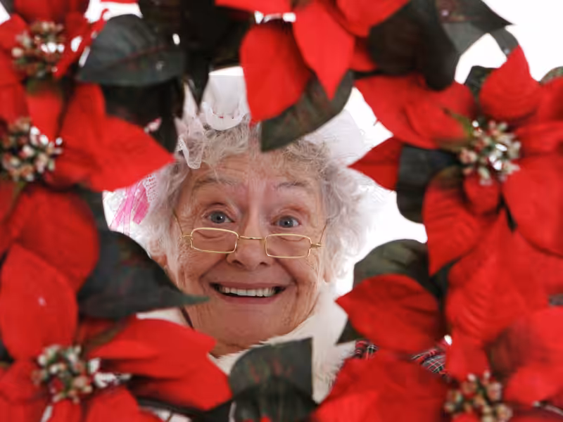 An elderly woman with glasses and a joyful smile is framed by a wreath of bright red poinsettias, looking directly at the camera.
