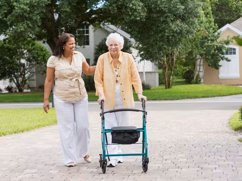 A younger woman supports an older woman using a walker as they walk together on a paved path outdoors, surrounded by green grass, trees, and houses in the background.