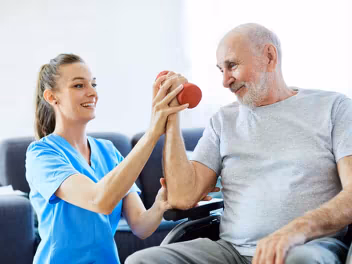 A young female caregiver in blue scrubs helps an elderly man in a gray shirt lift a small red dumbbell, both smiling and engaged in a rehabilitation or exercise activity indoors.