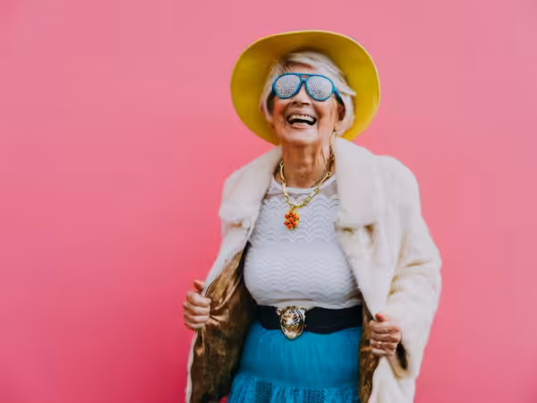 Elderly woman smiling, wearing a yellow hat, white fur coat, blue skirt, statement necklace, and large sunglasses with a polka-dot pattern, standing against a bright pink background.