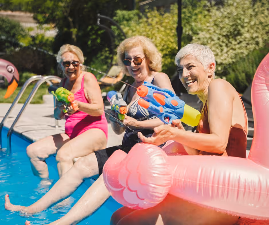 Three older women sit by a pool, laughing and spraying each other with colorful water guns. One is on a pink flamingo float. They appear to be enjoying a sunny day outdoors.