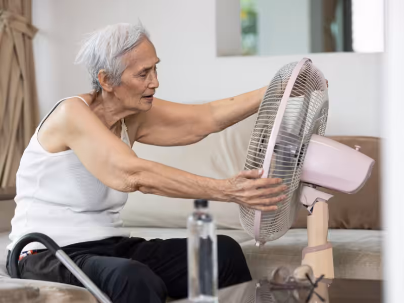 An elderly person with short gray hair sits on a couch, adjusting a large electric fan. The person is wearing a white sleeveless shirt and black pants. A water bottle is on the table in front of them.
