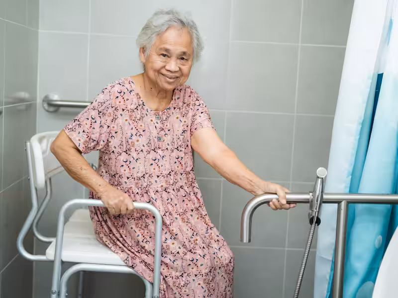Elderly woman in a pink patterned dress sitting on a shower chair in a tiled bathroom, holding onto metal grab bars for support and smiling at the camera.