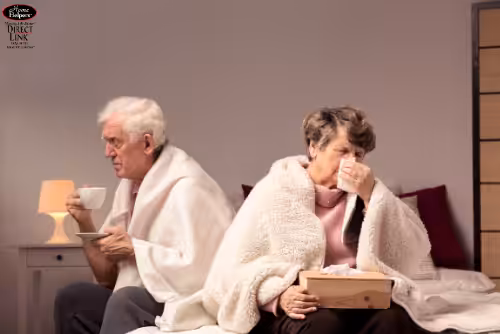 An elderly man and woman sit back-to-back on a bed, both covered in blankets. The woman holds a tissue to her nose, while the man sips from a white mug. A tissue box sits beside them, suggesting they are unwell.