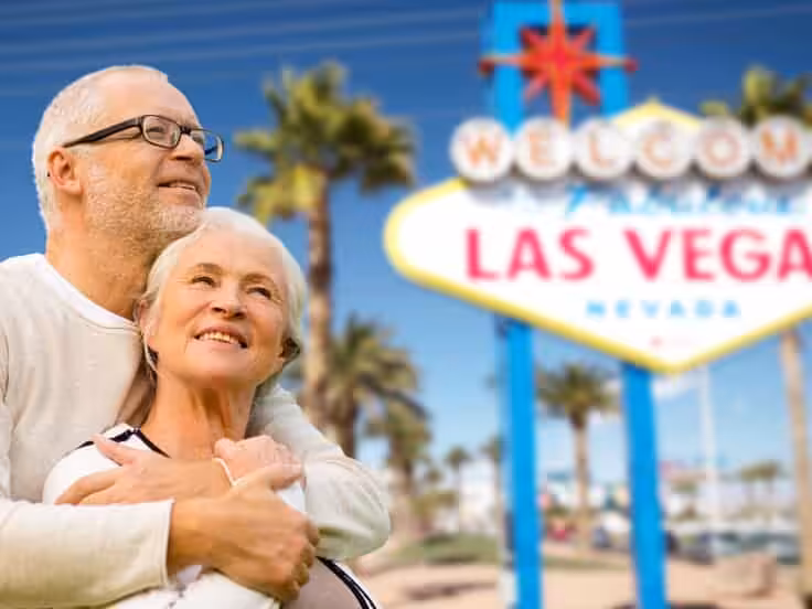 An older couple smiles and embraces in front of the famous Welcome to Fabulous Las Vegas, Nevada sign, with palm trees and a bright blue sky in the background.