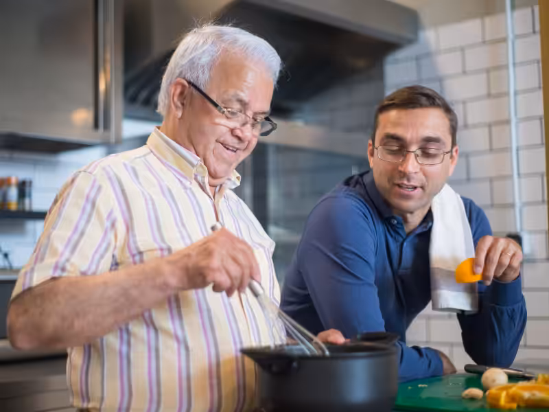 Two men cooking together in a kitchen. The older man is stirring food in a pot, smiling, while the younger man watches with interest, a towel over his shoulder and vegetables on the counter nearby.
