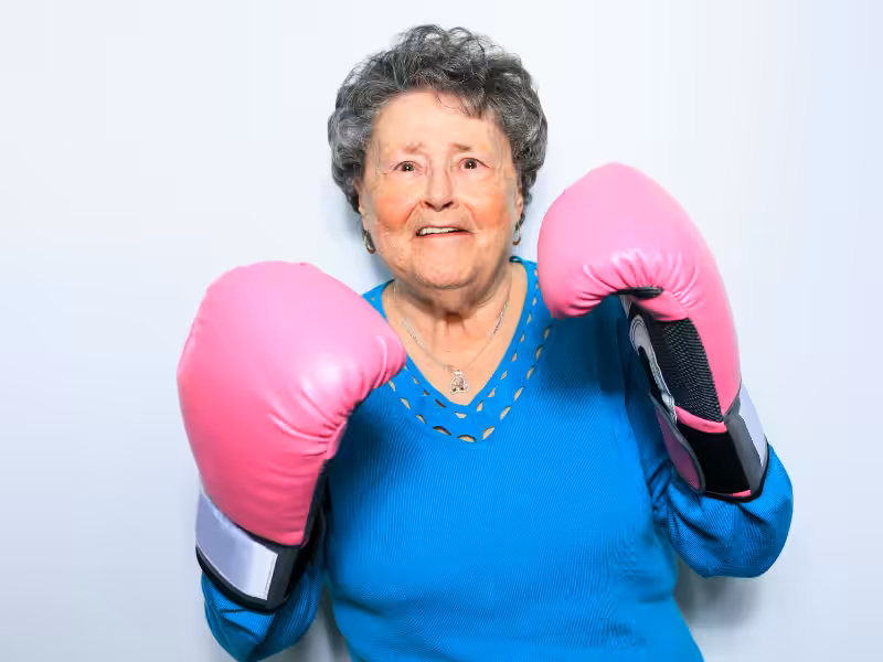 Elderly woman wearing a blue sweater and large pink boxing gloves stands in front of a white background, smiling and raising her fists in a boxing pose.