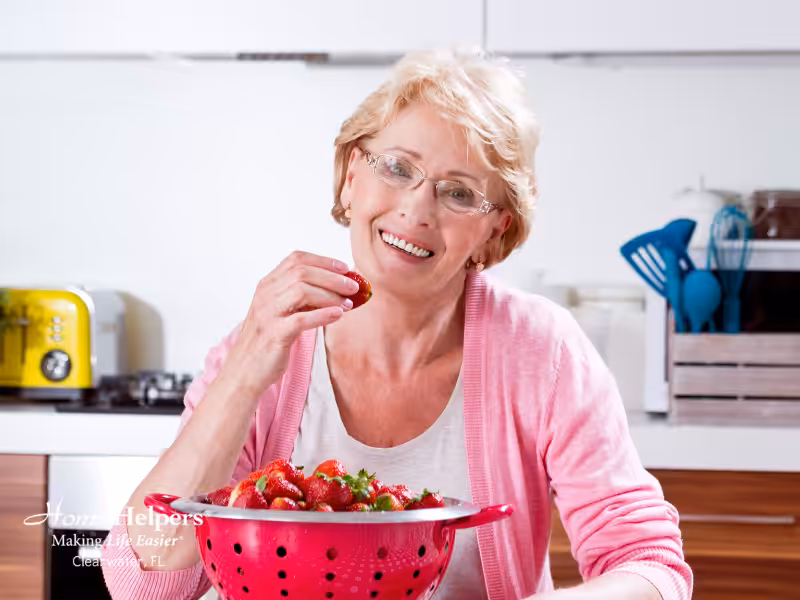 An older woman with short blonde hair and glasses smiles while holding a strawberry. She sits at a kitchen counter with a pink colander full of strawberries. Kitchen utensils and appliances are visible in the background.
