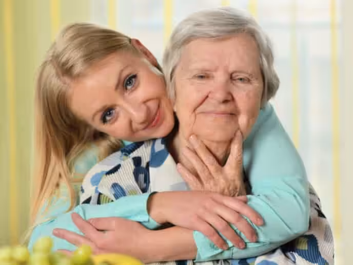 A smiling younger woman hugs an older woman from behind, both looking content. The older woman has gray hair and wears a patterned top, while the younger has blonde hair and wears a light blue shirt.