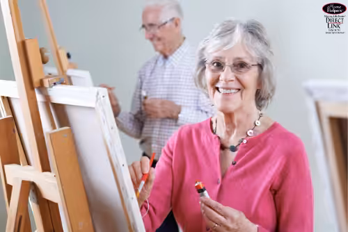 Two older adults painting at easels in a bright room. The woman in the foreground smiles at the camera, holding a paintbrush, while the man in the background paints on his canvas. Both appear happy and engaged in their activity.