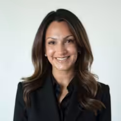 A woman with long, dark hair is smiling and wearing a black blazer over a black top. She is posed against a plain, light background.