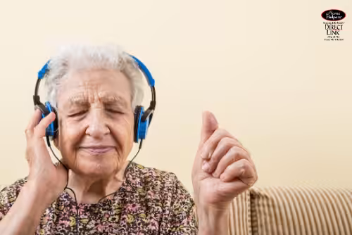 An elderly woman with gray hair wearing blue headphones sits on a striped couch, eyes closed and smiling, raising her hand as if enjoying music.