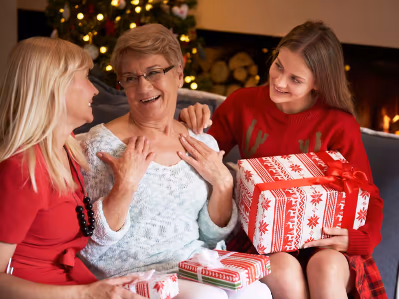 Three women sit together on a couch, smiling and holding wrapped Christmas gifts. A decorated Christmas tree and a lit fireplace are visible in the background, creating a warm, festive atmosphere.