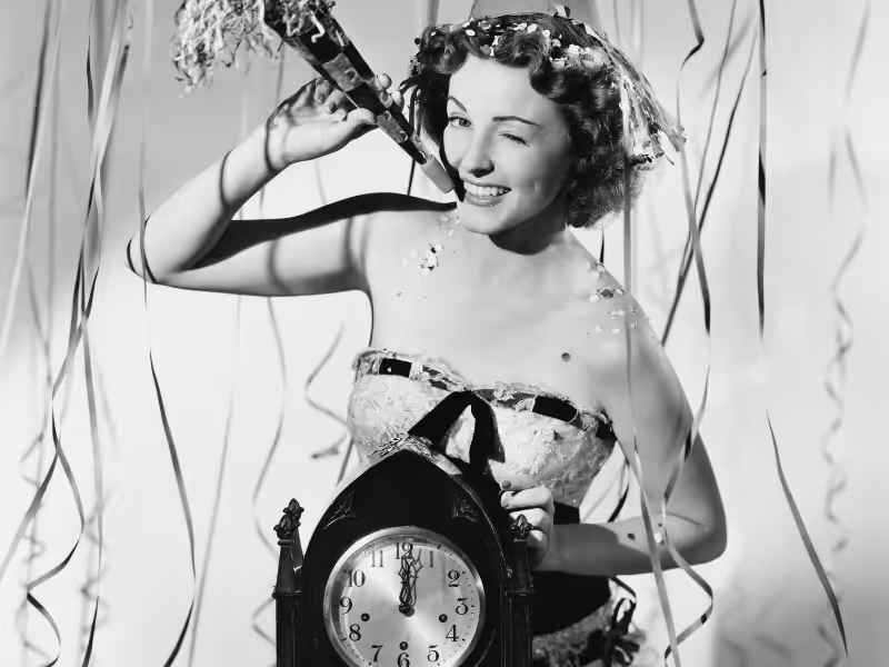 A woman in festive attire celebrates surrounded by streamers, holding a noisemaker and winking. She stands behind a clock showing midnight, suggesting a New Year’s celebration.