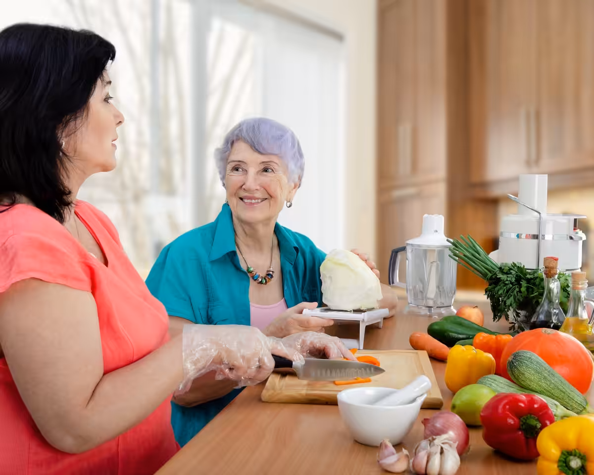 Two women preparing food in a kitchen together. One woman is slicing a carrot while the other, an older woman with gray hair, smiles and holds a cabbage. Various fresh vegetables are spread out on the counter.