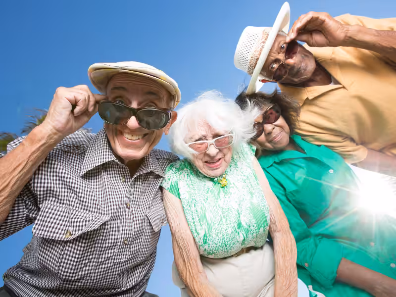 Four older adults are standing outdoors under a bright blue sky, smiling and playfully lowering their sunglasses while looking down at the camera. They are dressed in casual, colorful clothing and appear to be enjoying a sunny day.