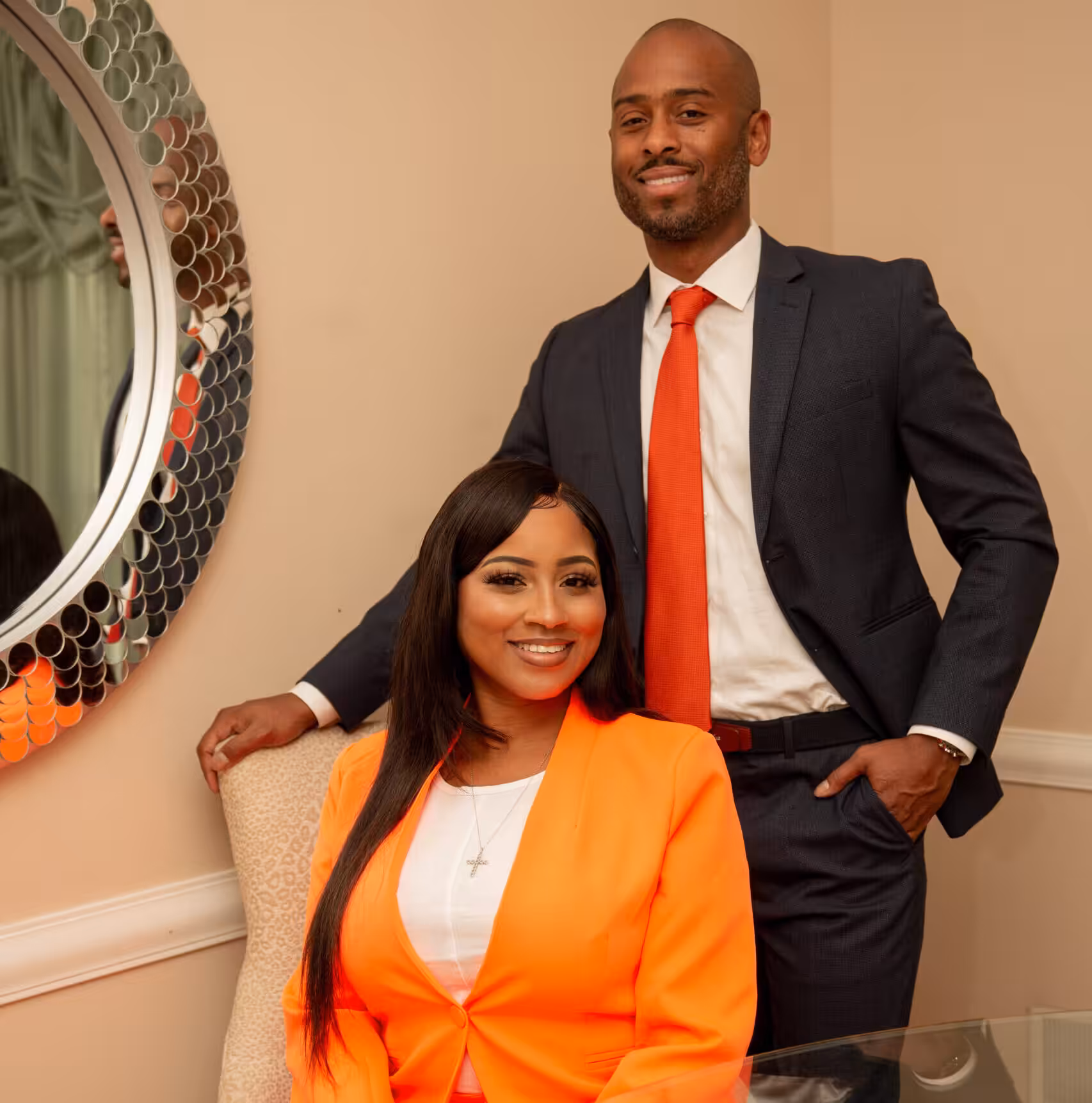 A woman in an orange blazer sits smiling at a table while a man in a dark suit and orange tie stands beside her with his hand on the chair, both looking at the camera in a warmly lit room.