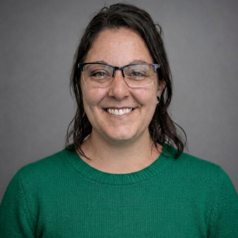 Person wearing glasses and a green sweater smiles in front of a plain gray background.