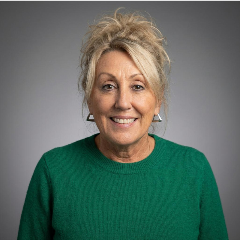 Smiling older woman with blonde hair in a casual updo, wearing a green sweater and triangular earrings, stands in front of a plain gray background.
