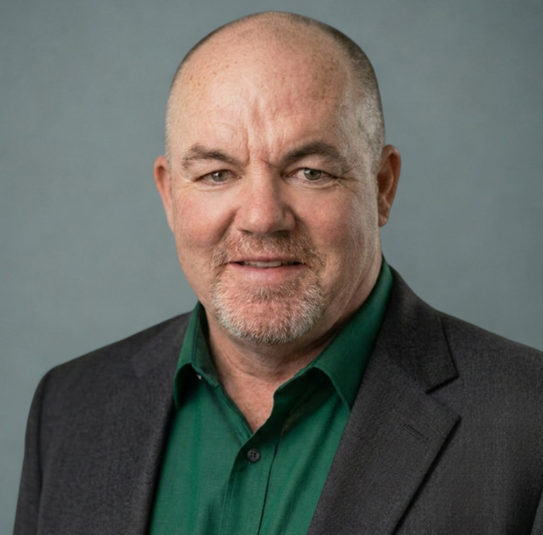 A middle-aged man with a shaved head, light facial hair, wearing a dark blazer over a green collared shirt, poses against a plain gray background.