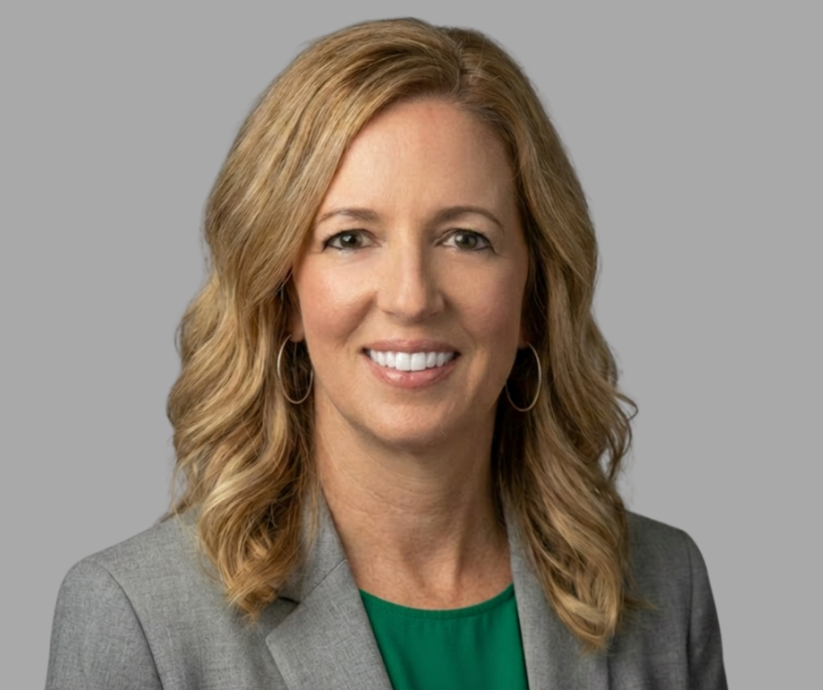 A woman with wavy blonde hair, wearing a gray blazer over a green top and hoop earrings, smiles at the camera against a plain gray background.