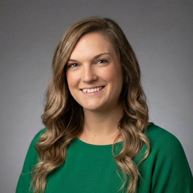 A woman with long wavy hair, wearing a green top, smiles at the camera against a plain gray background.