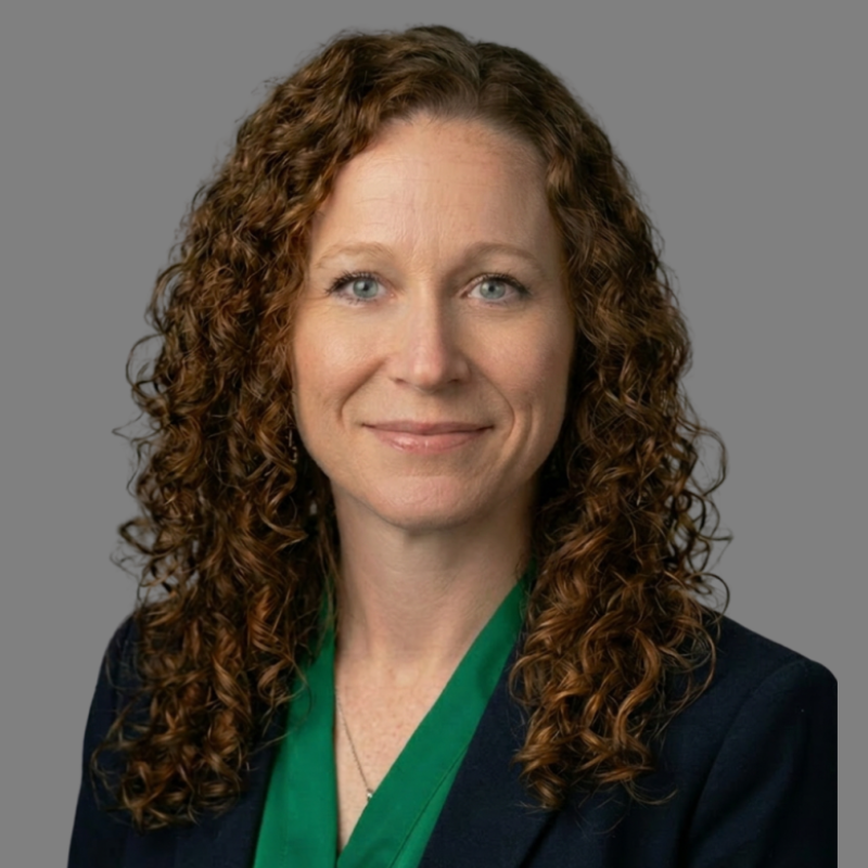 A woman with curly brown hair, wearing a green blouse and dark blazer, smiles slightly against a plain gray background.