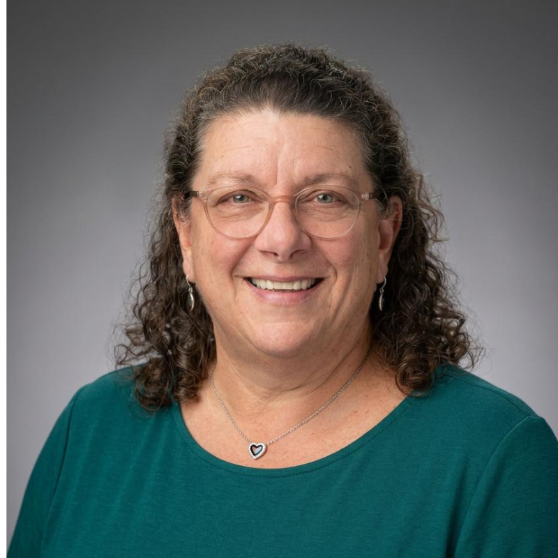 A woman with curly brown hair, wearing glasses, a teal shirt, and a heart-shaped necklace, smiles at the camera against a plain gray background.