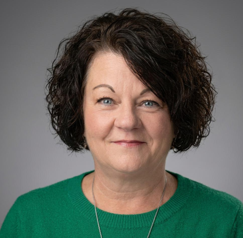 Woman with short, curly dark hair and blue eyes, wearing a green sweater and a silver necklace, smiling slightly in front of a neutral gray background.