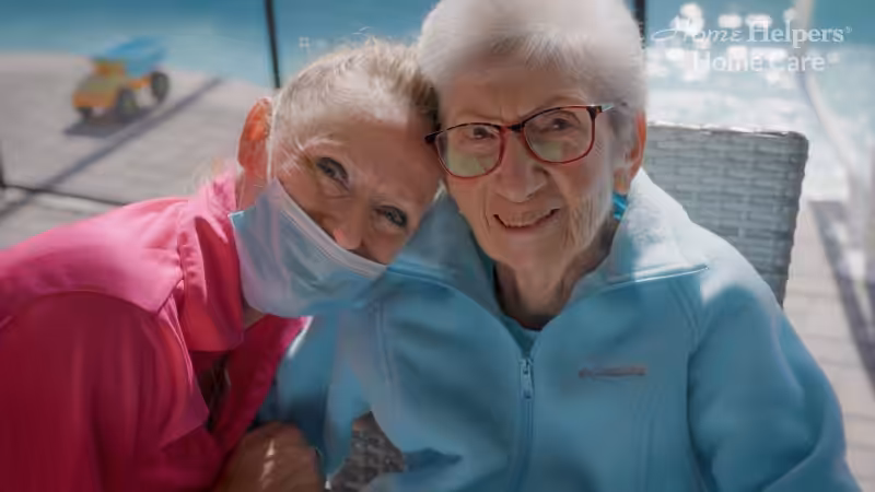A smiling elderly woman with white hair and glasses sits outdoors next to a caregiver in a pink shirt and face mask who is leaning in affectionately, both appearing happy and relaxed. The Home Helpers Home Care logo is in the corner.