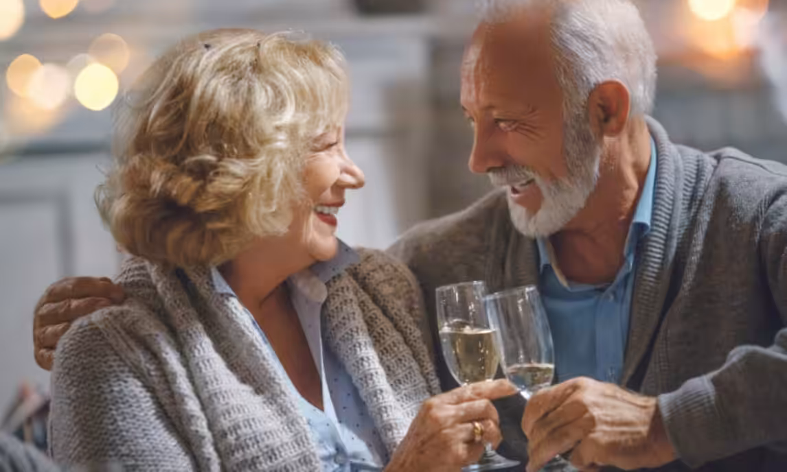 An older couple sits close together, smiling and looking at each other while clinking glasses of white wine in a warmly lit setting. Both wear cozy sweaters, creating a joyful and intimate atmosphere.