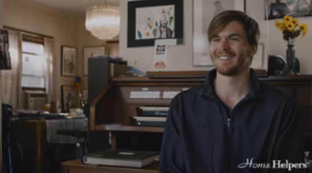 A smiling man with light brown hair and a beard sits in a cozy room with art on the walls, a piano behind him, and the Home Helpers logo in the bottom right corner. A vase of yellow flowers is on the piano.