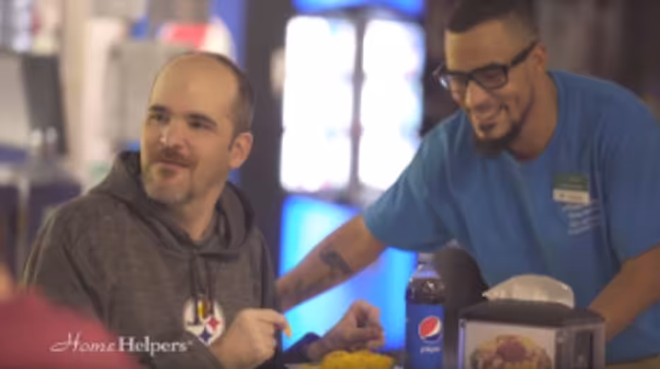 A man wearing a Pittsburgh Steelers hoodie sits at a table with food, smiling, while another man in a blue shirt and glasses stands beside him, also smiling. A Pepsi bottle and a napkin holder are on the table.